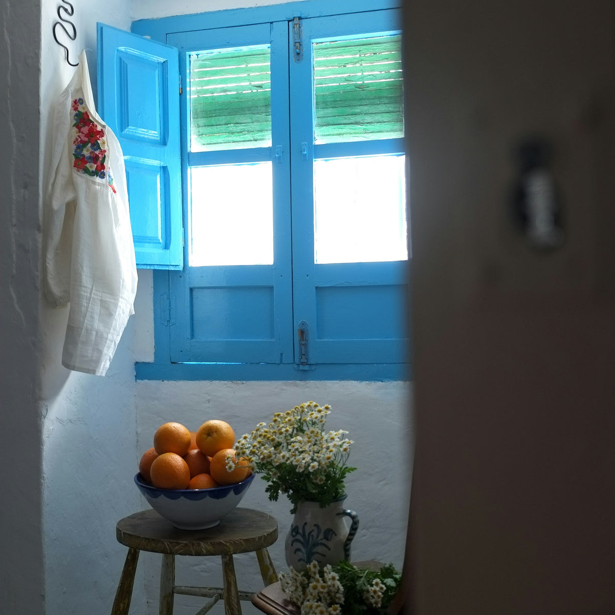 Rincón encalado con contraventanas azules, cuenco de naranjas y flores en jarra de cerámica
