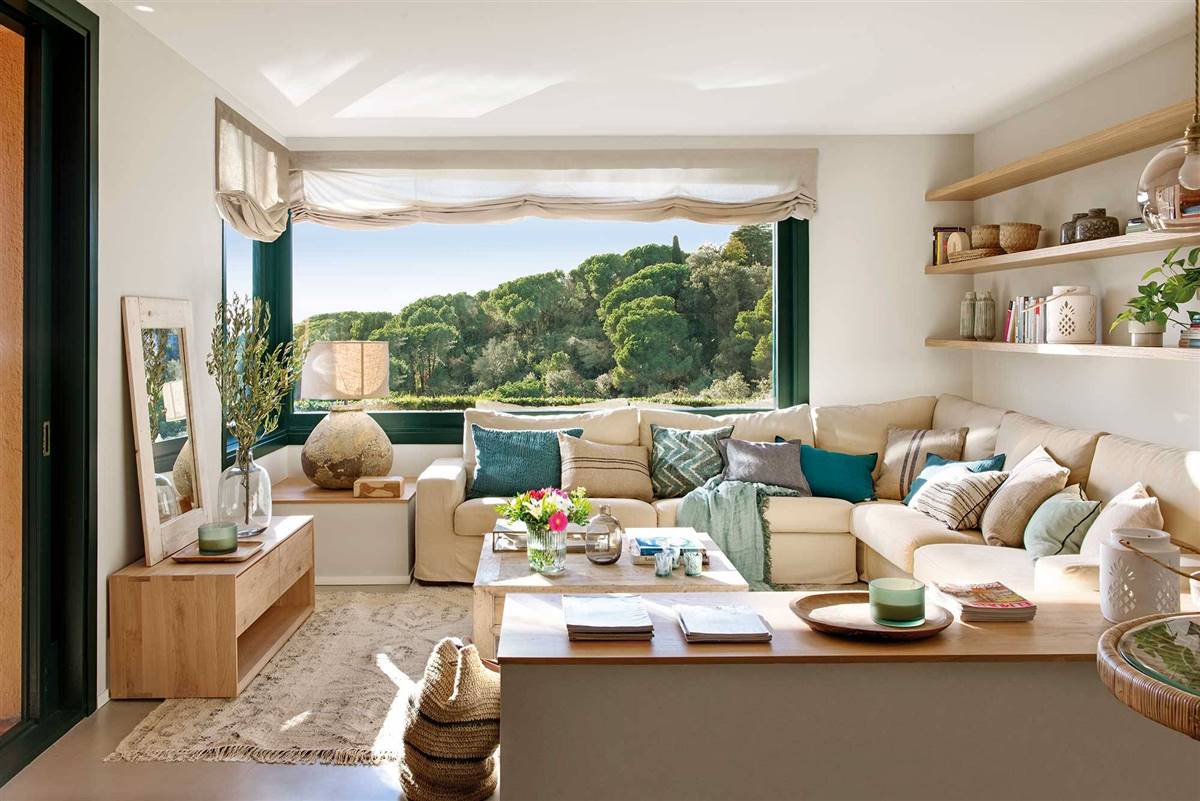 Salón con sofá rinconero claro lleno de cojines frente a un gran ventanal con vistas a un pinar