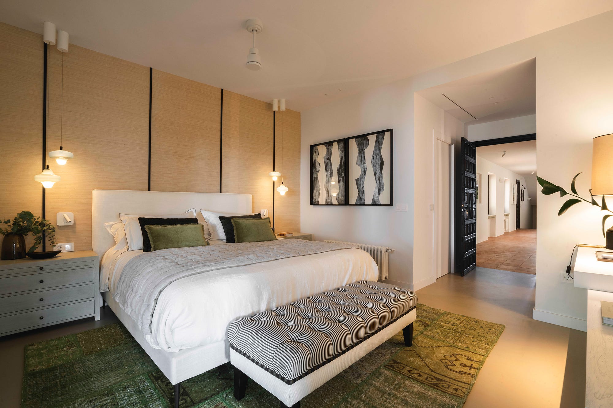 Habitación de invitados con cama de ropa blanca, cojines verdes, banco tapizado de rayas a los pies y lámparas colgantes