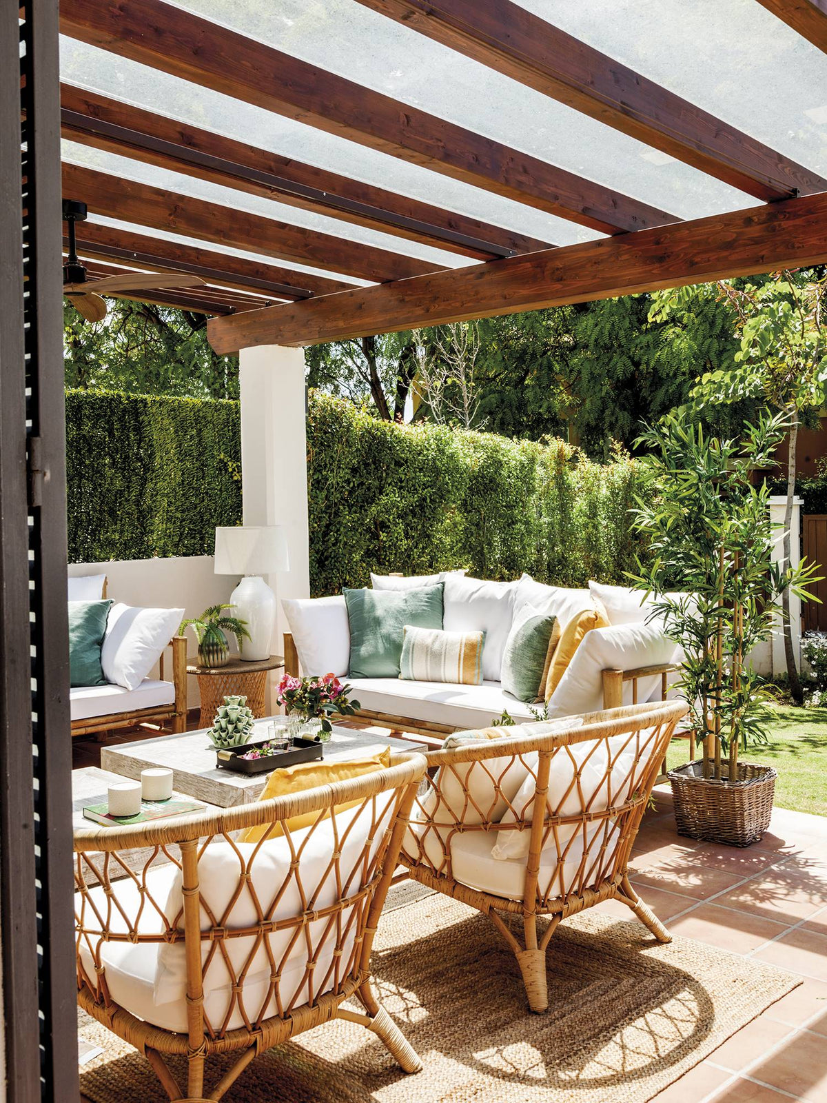Porche mediterráneo bajo una pérgola de madera con sofá blanco, cojines en tonos suaves y sillones de fibra